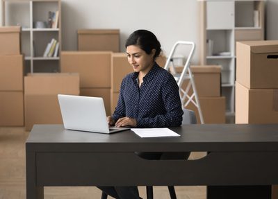 woman sitting on a desk on a laptop and boxes in the background