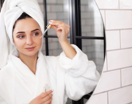 A woman wearing a bathrobe uses a face serum and examines her reflection in the mirror
