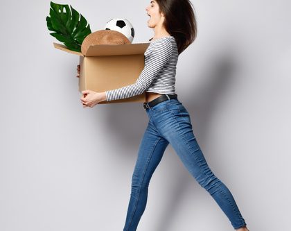 woman jumping and holding a cardboard box containing various items including a plant and a soccer ball