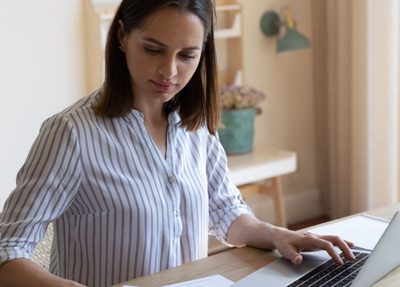a woman sits at a table, focused on her laptop with calculator