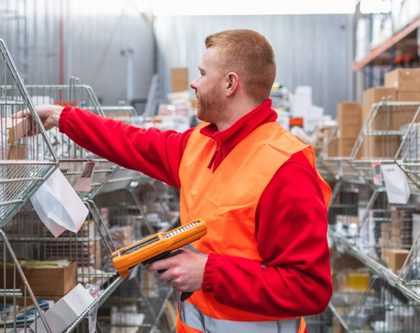 a man in a warehouse wears an orange vest and holds a clipboard